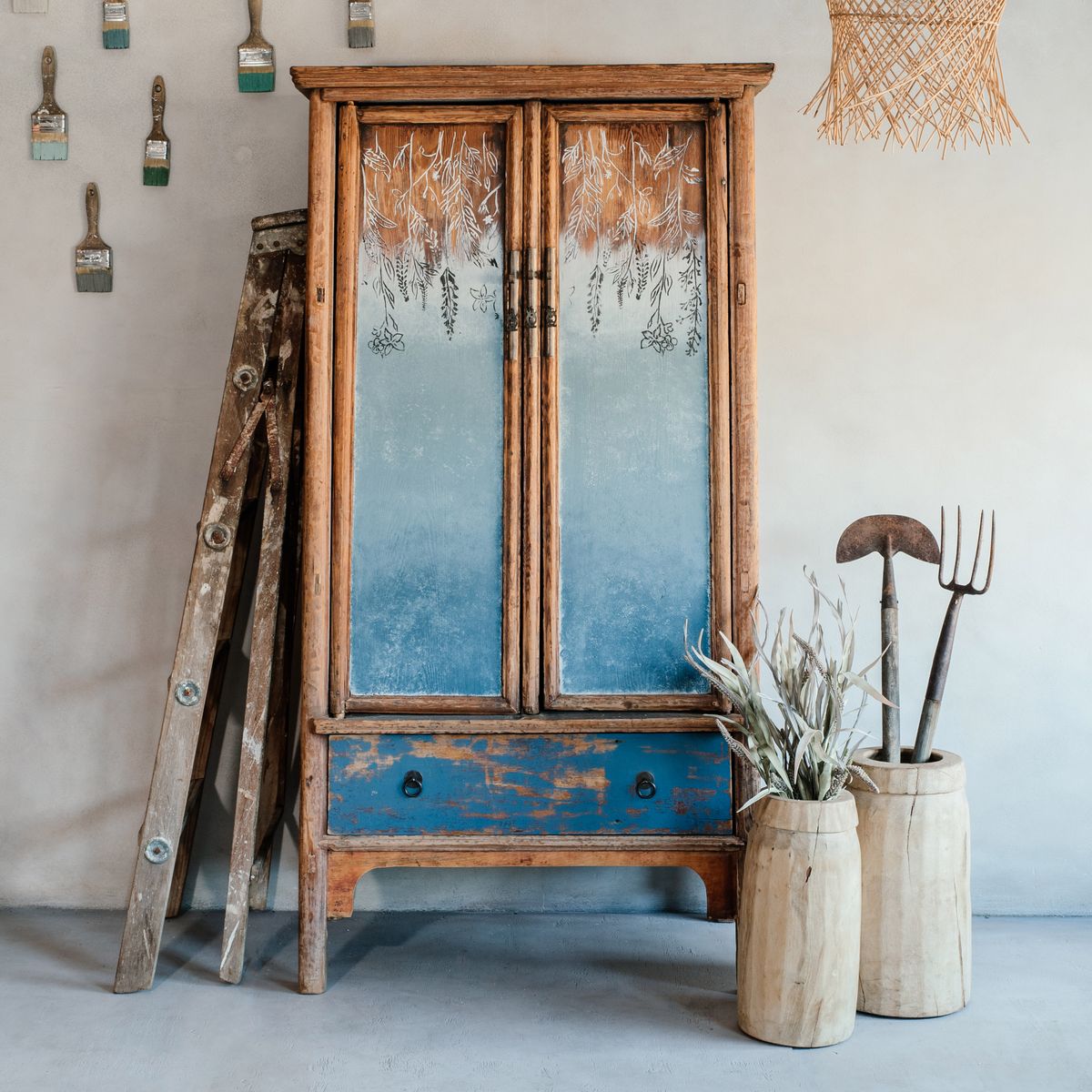 A bucket of Godfrey's Glimpse wall paint in a moody blue-grey color, displayed in a rustic wooden setting with a old-fashioned mirrored cabinet and decorative forks.