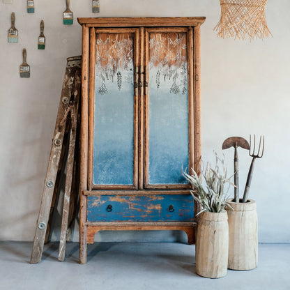A bucket of Godfrey's Glimpse wall paint in a moody blue-grey color, displayed in a rustic wooden setting with a old-fashioned mirrored cabinet and decorative forks.